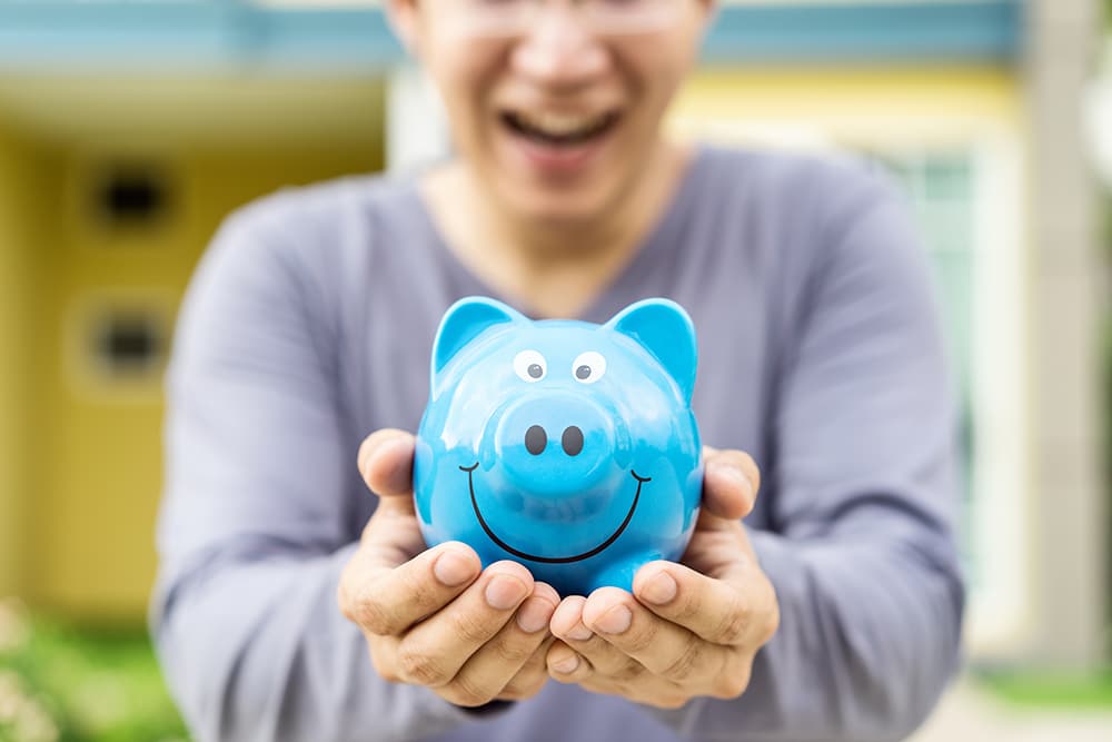 Happy asian man holding blue piggybank with front outdoor home b